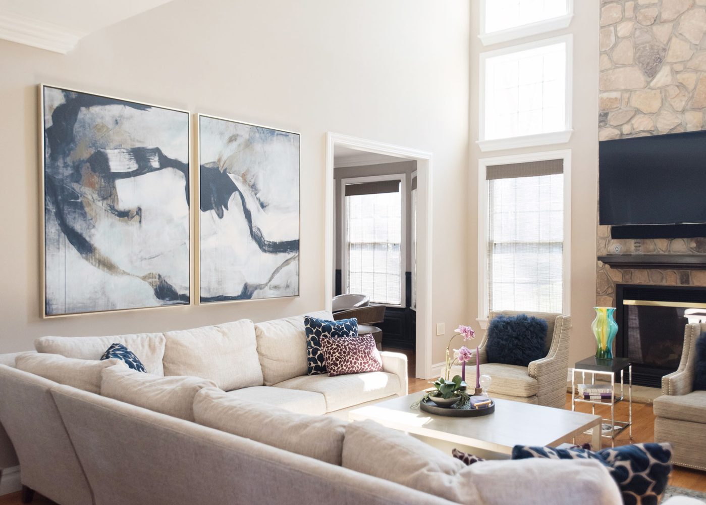 Family room with pale beige walls and matching sectional sofa. Wall has two large abstract artworks with navy, beige and white shapes. Tall stone fireplace with two beige upholstered arm chairs in front.