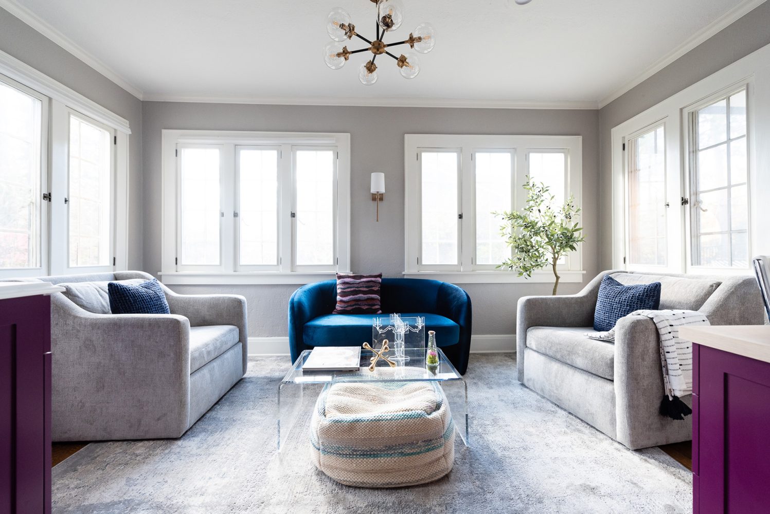 Family room surrounded by windows. Walls are painted medium gray. A blue velvet loveseat is flanked by two gray sofas. There is a clear glass coffee table. The edges of purple cabinets are seen on either side of the image.