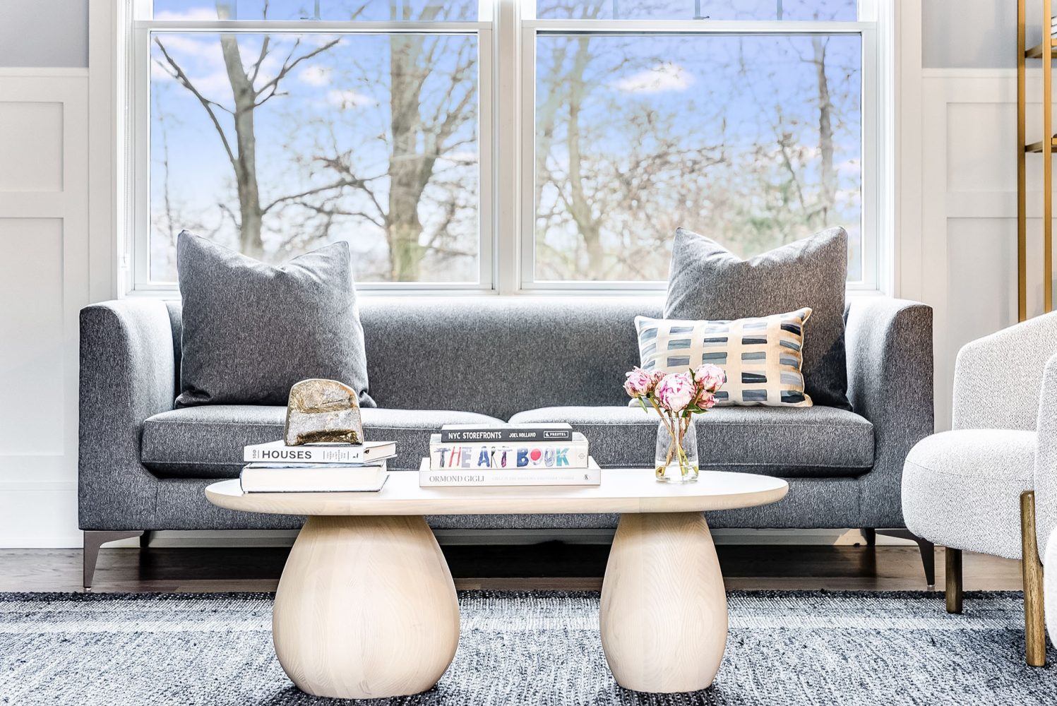 A modern light-finished wooden coffee table with bulbous legs is shown in front of a grey tweed sofa and large picture window.