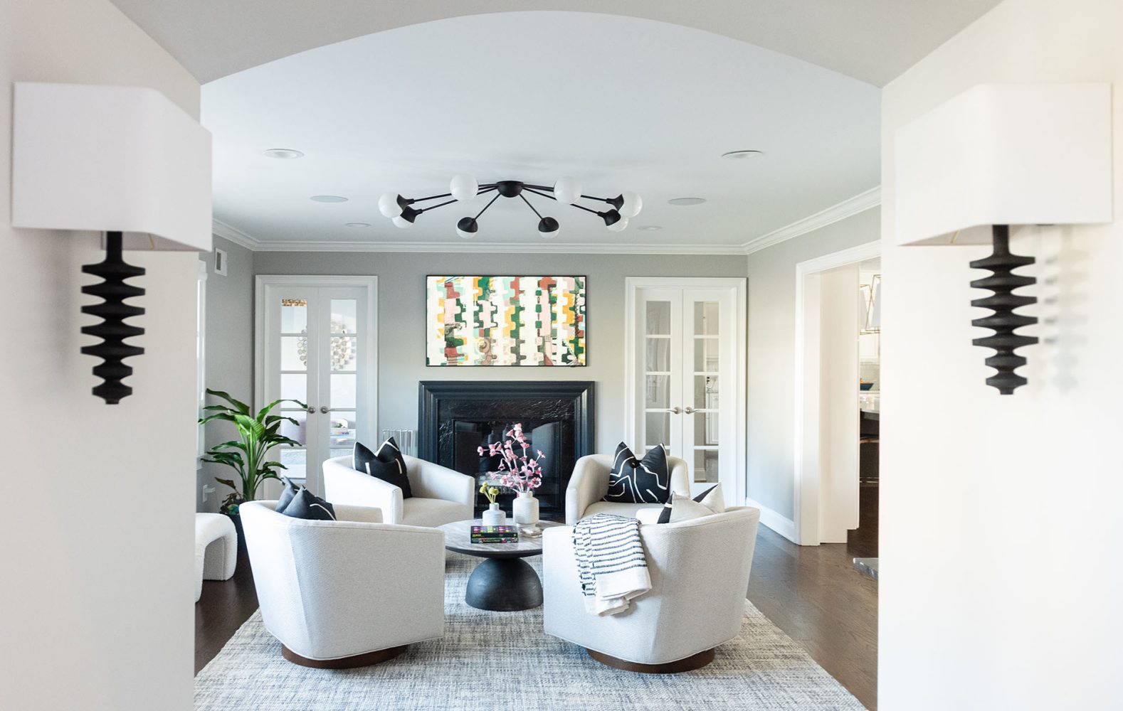 View from hall into living room. Hall walls feature sconces with abstract tiered black bases and rectangular white shades. Living room has four white-upholstered tub chairs around a circular coffee table with marble top and steel base. Black fireplace and modern chandelier with black metal and white globes.