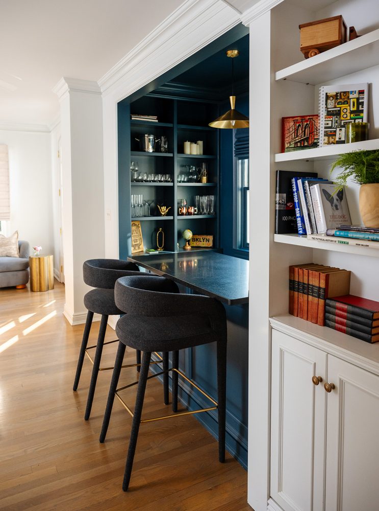 A bar with blue walls and counter and two upholstered barstools is built into a wall bordered by a white wooden bookcase.