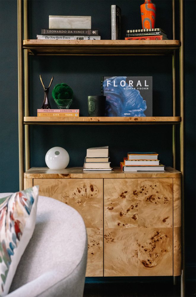 Tall bookcase with brass rail sides and burl shelves and lower drawers in front of a teal wall.