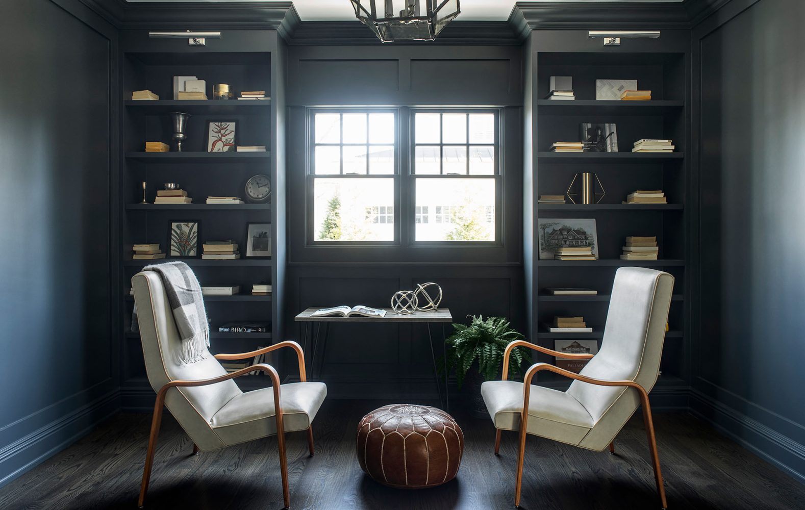 A room with walls and bookcases painted dark blue-gray. The layout is symmetrical with one bookcase on left, double window in middle, and one bookcase on right. Bookcase shelves are styled with books and objects and two white modern chairs are placed in front.