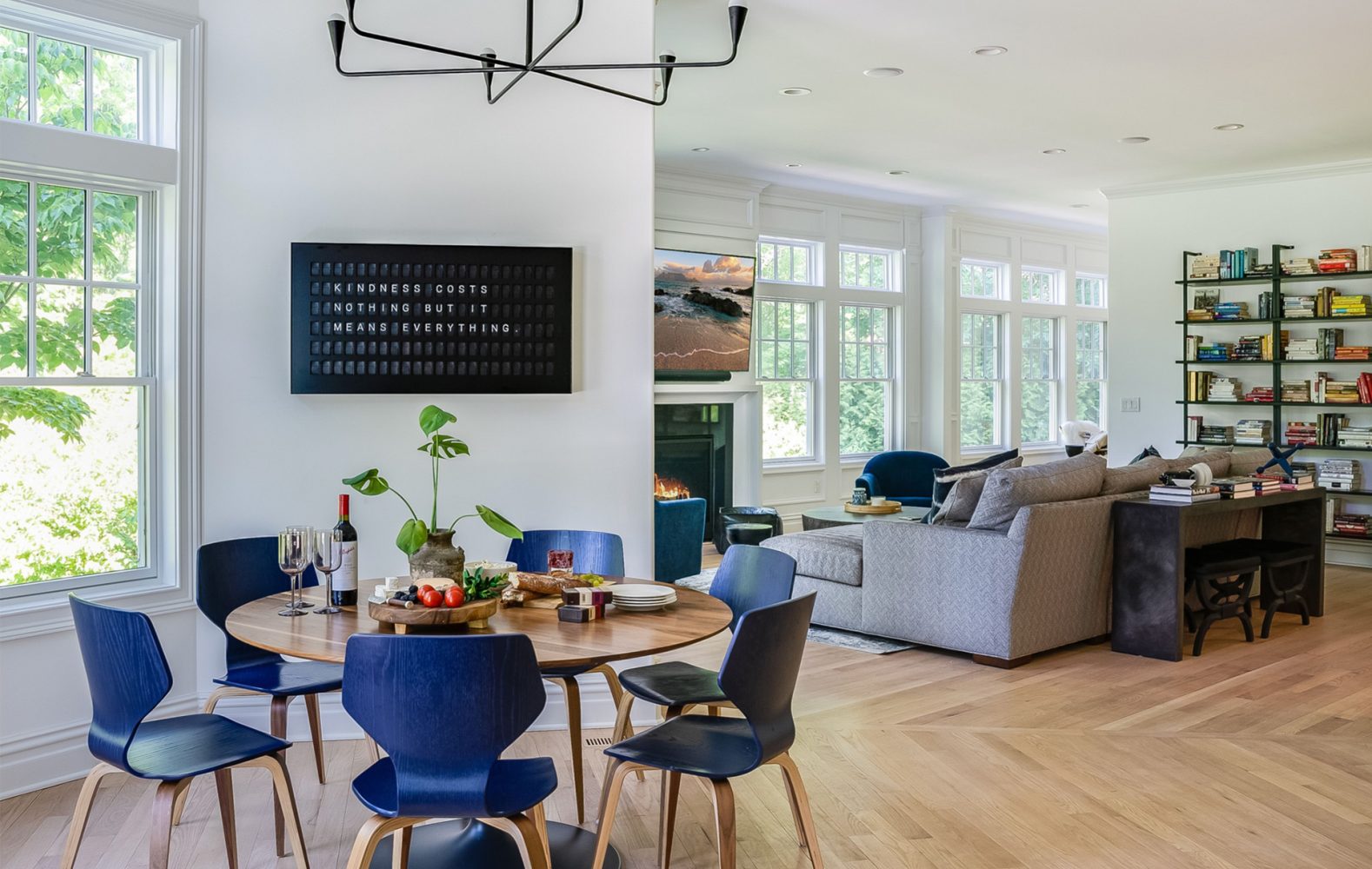 Large room with white walls and herringbone light wood floors. Round table has blue-stained wood chairs around it. There is a vestaboard on the wall above the table. In the background is a gray couch, large TV, and bookshelf.