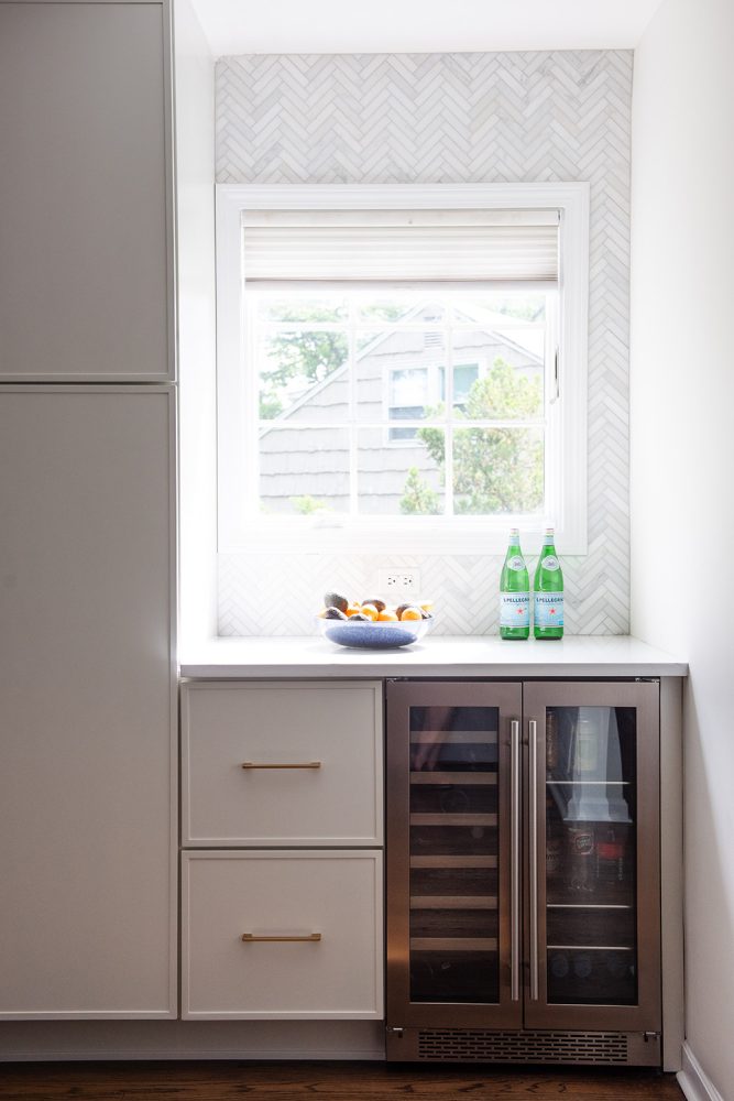 Built-in cabinetry beneath a window that has two square drawers and a stainless wine fridge. Countertop above is white. Window surround is white marble tile in a herringbone pattern.