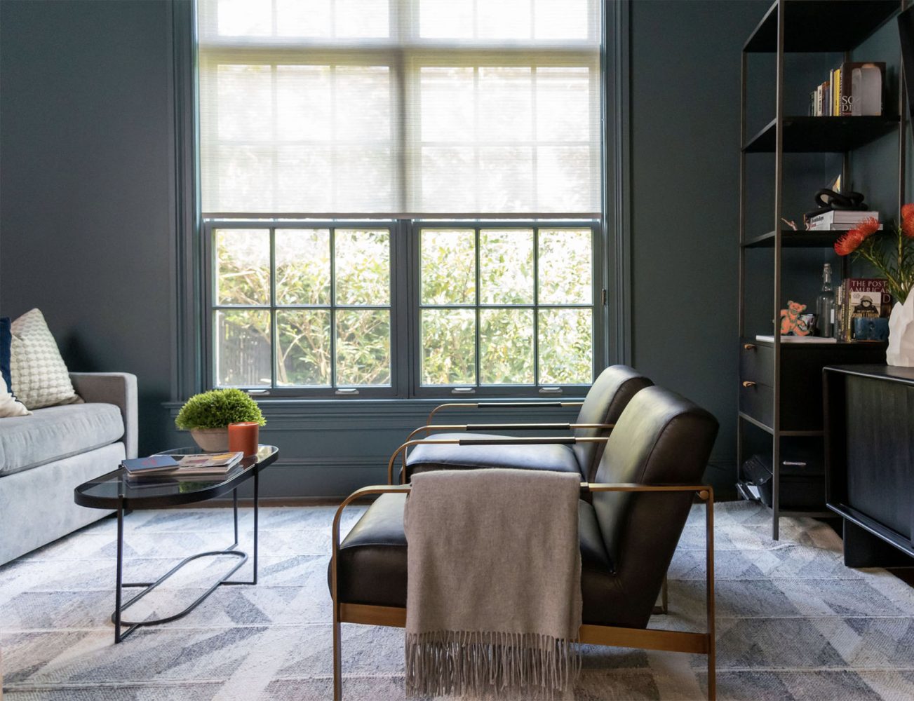 An office with medium gray painted walls, two modern club chairs with brass arms and black leather upholstery, a brown metal and glass cocktail table, and minimalist black bookcases.