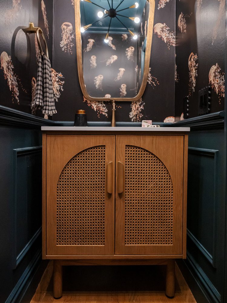 Closeup of a medium-wood vanity with arched caned inset, aged brass faucet and mirror, and walls with pink jellyfish against a black ground.
