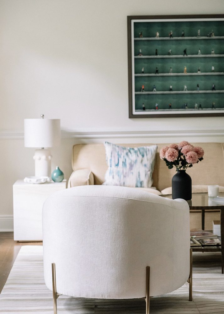 A living room featuring a beige sofa, off-white upholstered side chair, a white side table and lamp, and a large artwork above the sofa that features a grid of people walking in front of a dark green wall.