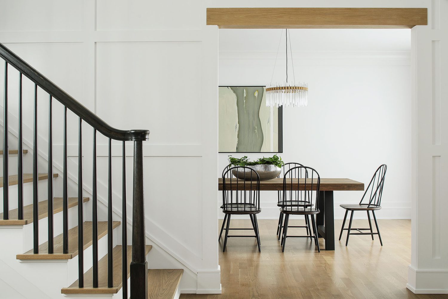 View from a black-railed staircase into a white-walled dining room with wood floors, black chairs, and an off-white and grey abstract piece of art hung over a wooden dining room table.