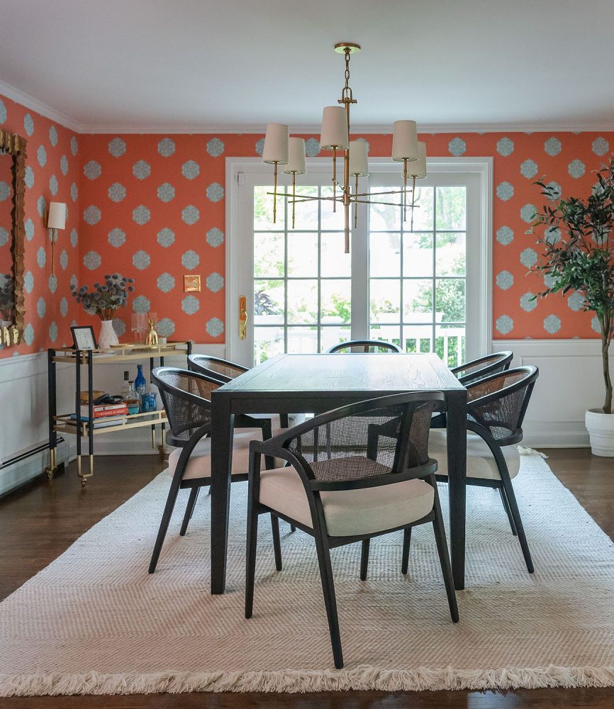 Dining room with salmon-colored wallpaper strewn with white and grey medallions. There is a transitional brass spoked chandelier above a dark wood rectangular table. The chairs are dark wood with caned backs and upholstered seats.