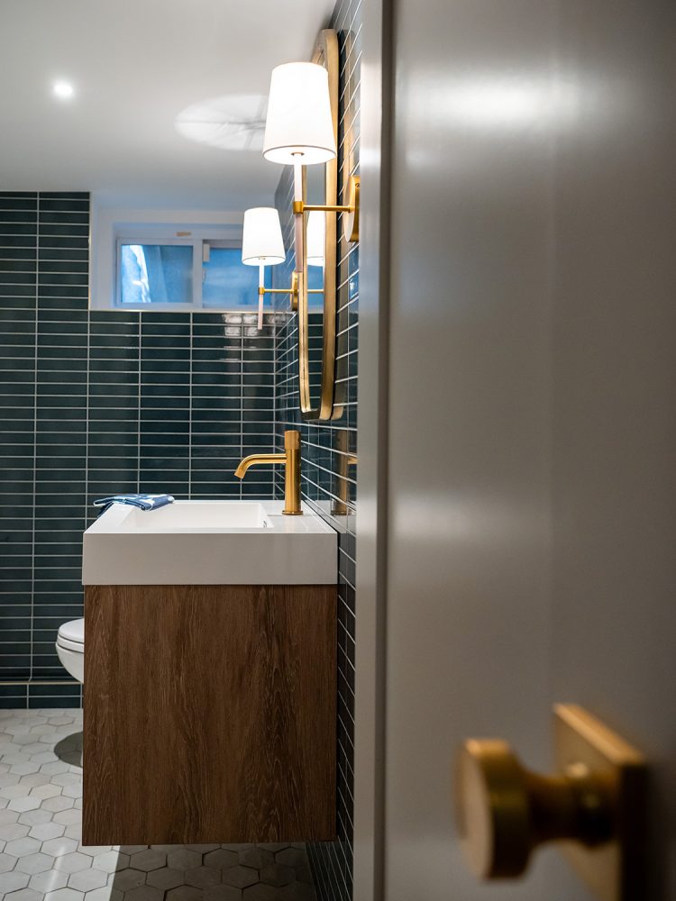 A bathroom with a grid of blue rectangular subway tiles on the walls, modern brass mirror, faucet, and sconces, and floating medium wood vanity.