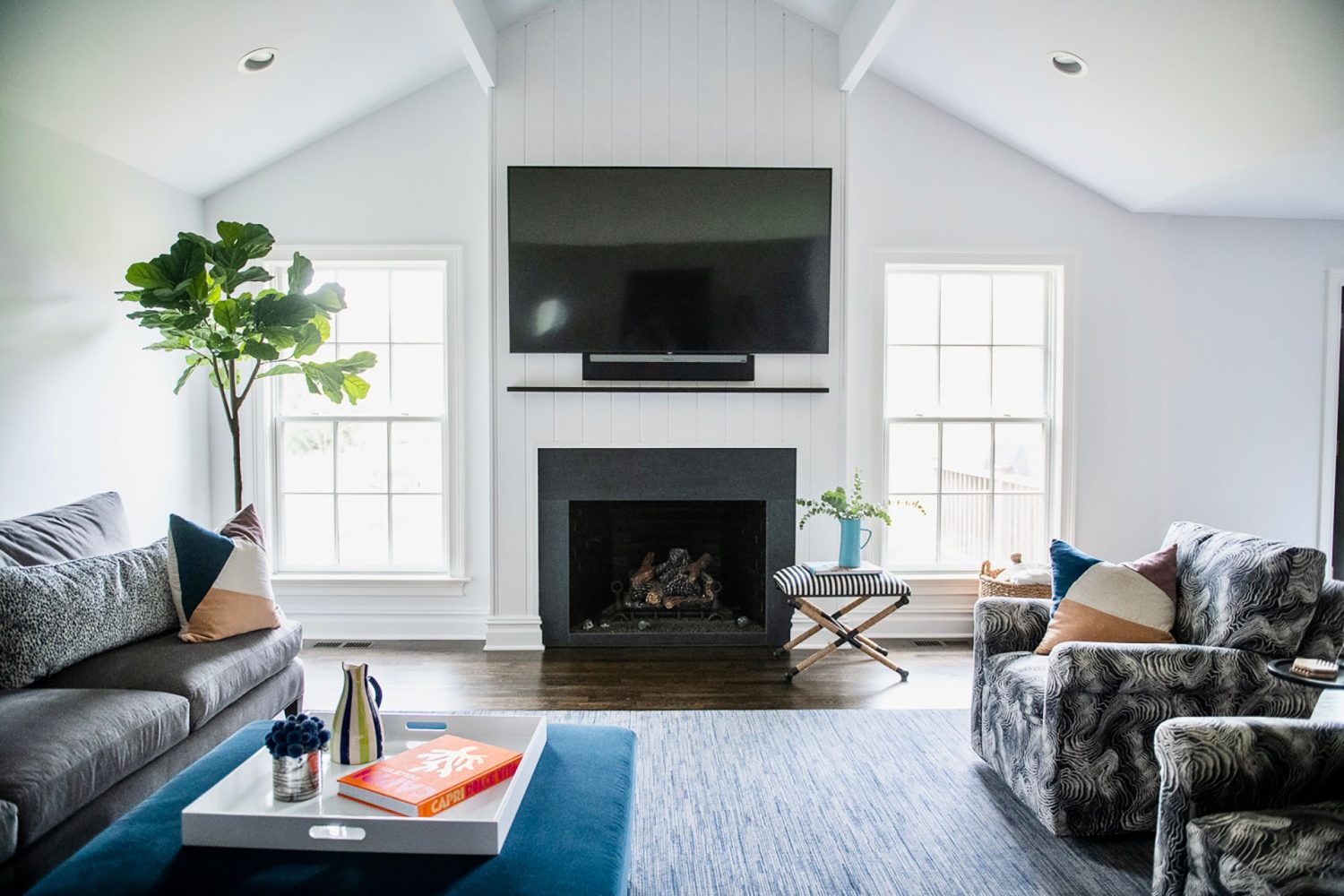 Living room with tall fireplace encased with white vertical boards. Surround of fireplace is dark grey stone. Large TV is mounted above fireplace, left wall has grey upholstered couch, and right wall has two patterned easy chairs.