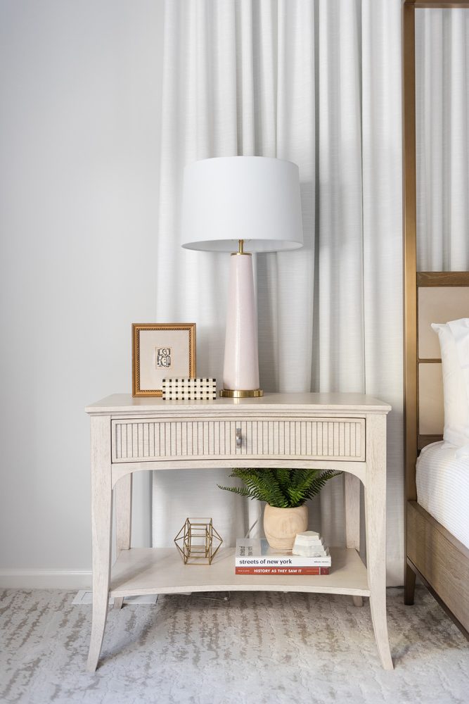 A weathered white nightstand with fluted drawer is topped with a slender brass and light pink lamp. Nightstand is styled with a small brass and linen framed picture, a small fern, and other books and objects.