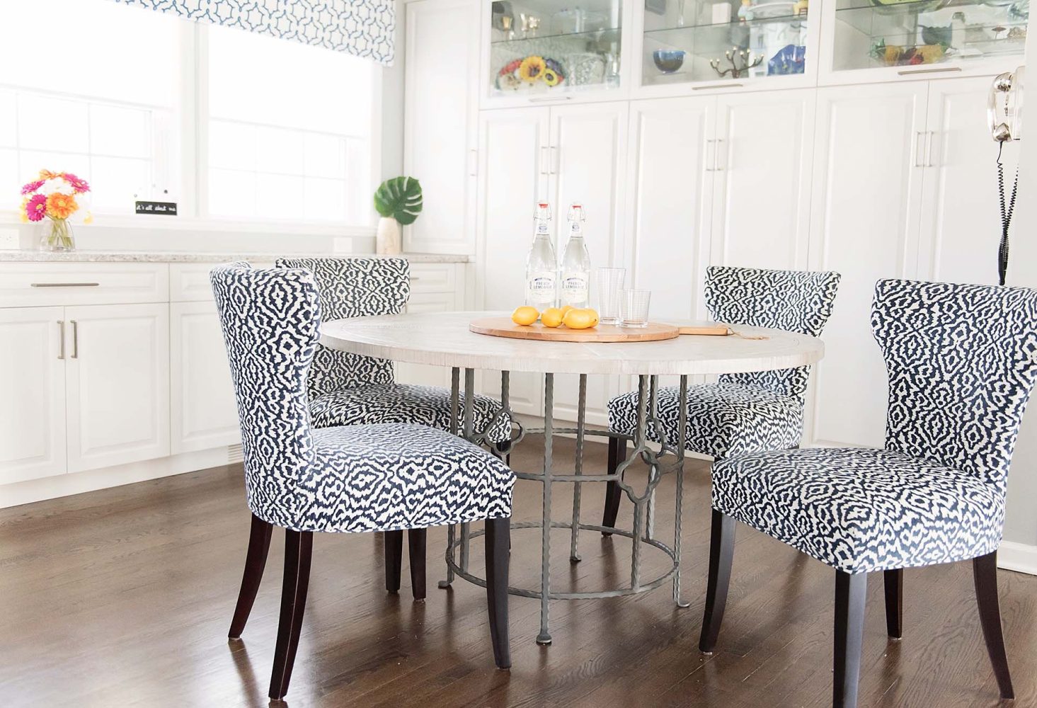 White built-in cabinetry along back wall; cabinets have upper glass shelves and doors. Round table with four upholstered chairs in navy blue and white pattern.