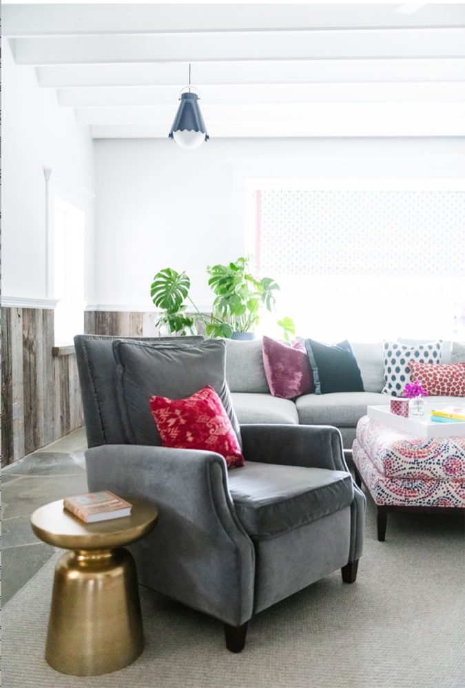 Family room with white walls and grey wood wainscotting. Floor is grey stone with light grey area rug. Grey velvet easy chair in foreground with brass occasional table. Light grey sofa in background, with ottoman upholstered in blue and berry dotted swirl print fabric. Throw pillows in blue and berry tones and prints.