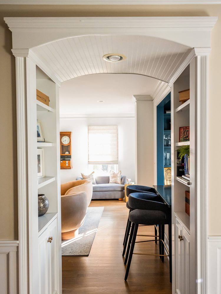 Arched, white painted wooden entranceway to a serene living room. Can catch a glimpse of plush seating in shades of blue, grey, and beige.