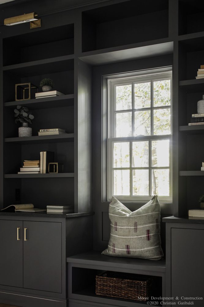 A close view of dark gray bookcases with a small window seat. Window seat has dark woven basket beneath and light gray striped pillow above. Bookcase is styled with small plants, antique metal objects, and books.