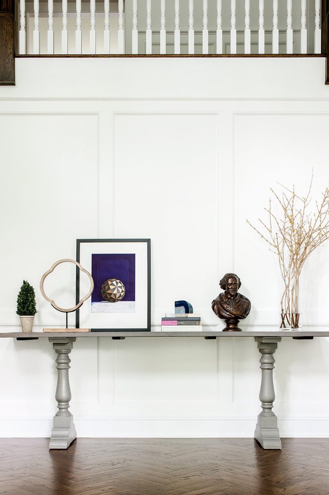 A grey wood table in a paneled, white walled foyer, with stair railing above and herringbone wood floor beneath. On the table is a small plant, several objets d' art, and books.