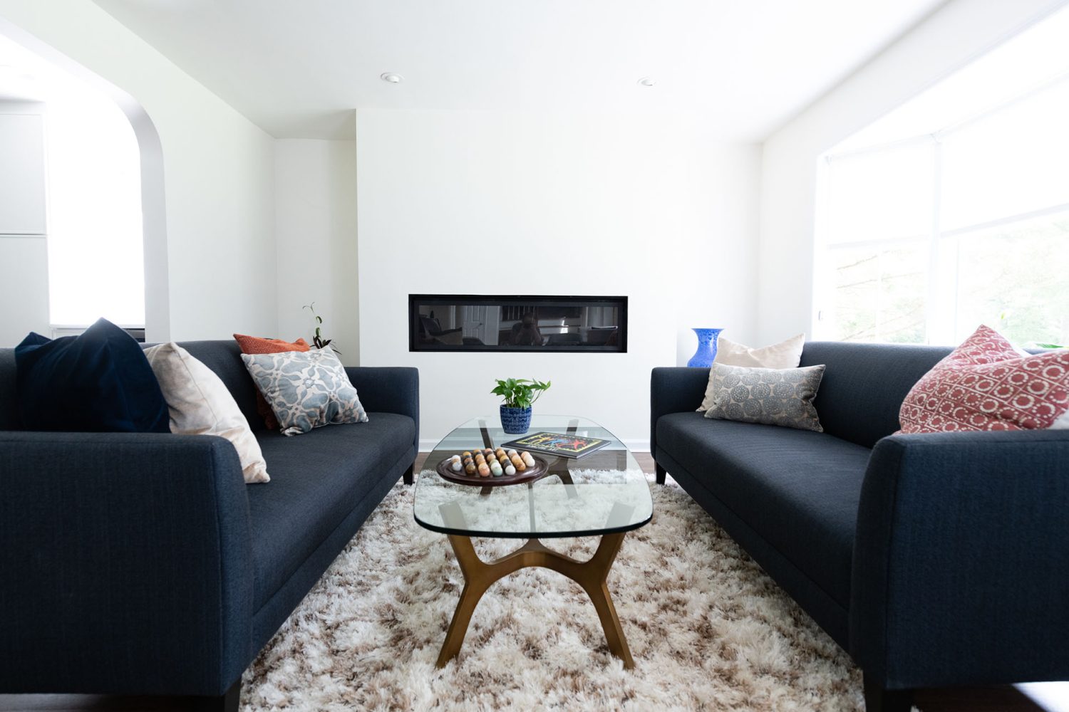 White-walled living room with two navy couches, a glass and wood coffee table, and deep shag rug. On the back wall is a modern, slim, inset black fireplace.