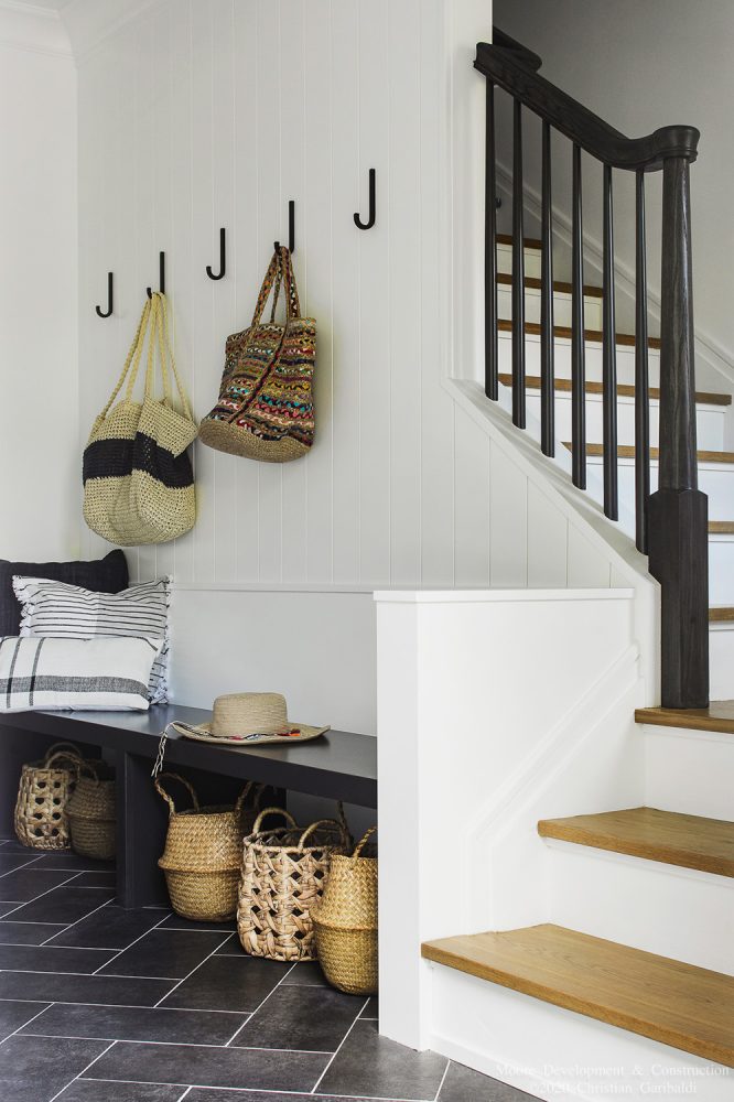 A black bench beneath a white-walled stairwell, staged with a variety of round baskets under the bench and woven bags hung on black hooks above.