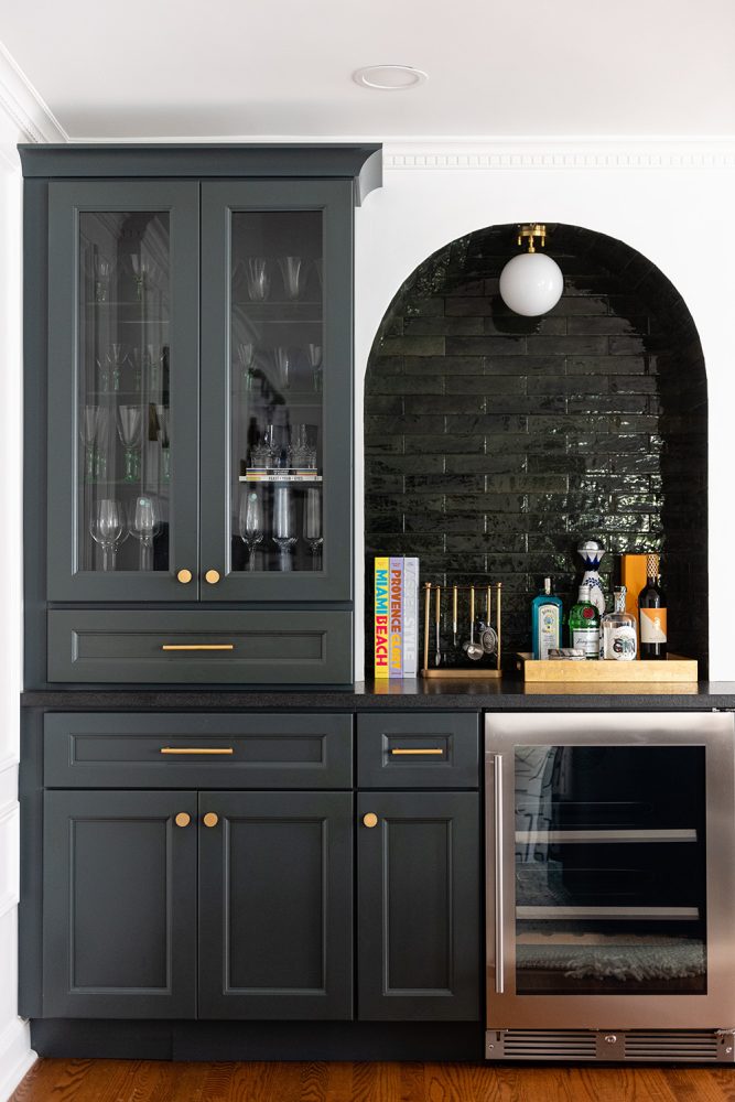 Built-in dark grey cabinetry with modern brass hardware and darker grey countertop. Stainless steel wine fridge beneath a bar that is a glossy black tiled wall alcove