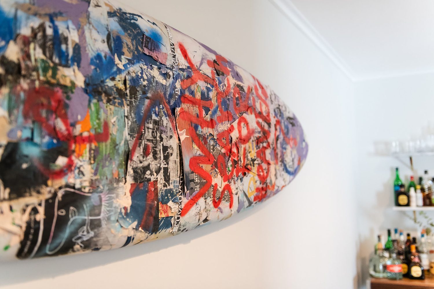 Close view of a sticker and graffiti-covered surfboard hung on a white wall near a bar.